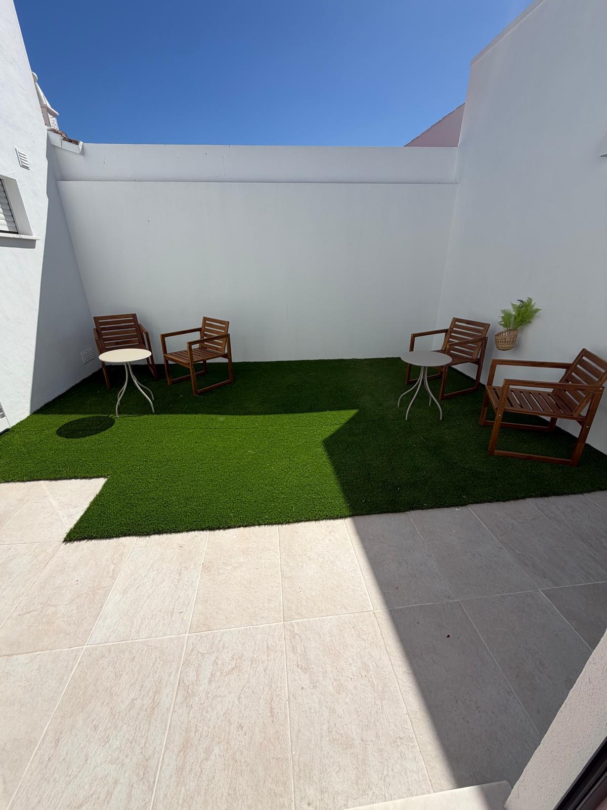 Pátio exterior com relvado, cadeiras de madeira e paredes brancas ao sol